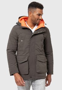Chaqueta acolchada verde oliva con capucha de color naranja brillante, que cuenta con dos bolsillos frontales y cierre de cremallera. Tela texturizada, diseño aislante.