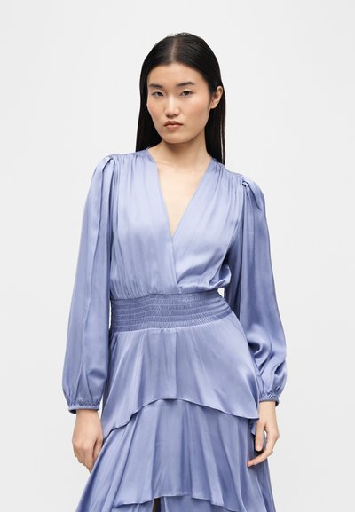 Νεαρή γυναίκα με μακριά σκούρα μαλλιά, φορώντας ανοιχτό μωβ σατέν φόρεμα με φουσκωτά μακριά μανίκια και στρωμένο φόρεμα, μπροστά σε ένα απλό φόντο.