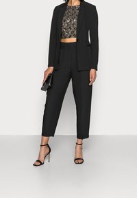 Blazer nero su un top cropped con paillettes, abbinato a pantaloni neri su misura e tacchi a schiocco neri. Include una piccola borsa a tracolla nera.