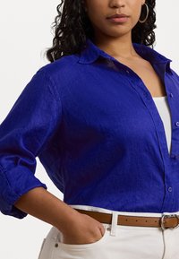 Camisa de linho azul royal com mangas arregaçadas, fecho à frente com botões e um corte descontraído, combinada com um cinto bege e calças brancas.