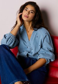 Šviesiai mėlyna ilgarankovė džinsinė palaidinė su giliais V formos iškirpimais ir juodais širdies formos papuošimais, derinama su plačiomis tamsiomis džinsinėmis kelnėmis. Sėdi priešais raudoną foną.