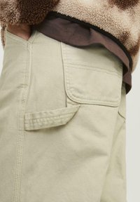 Beige cargo bukser har en sidelomme med klap, robust stoftekstur og synlige syninger. Båret med et brunt lag under.