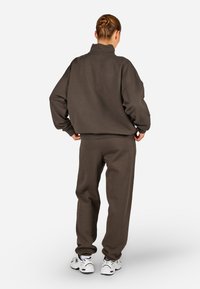 Brązowy, oversize'owy sweter oraz pasujące spodnie wykonane z miękkiego materiału, z żeberkowymi mankietami i wysokim kołnierzem, w połączeniu z białymi trampkami.