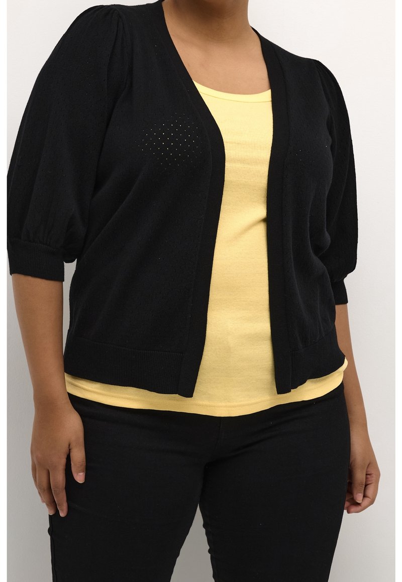 Personne portant un cardigan noir ouvert devant avec des manches bouffantes sur une chemise jaune et un pantalon noir, debout devant un fond uni.