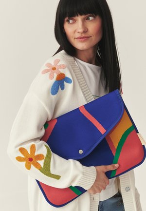 Žena s rovnými čiernymi vlasmi, oblečená v bielom kardigane s farebnými kvetinovými vzormi, drží veľkú modro-červeno vzorovanú listovú kabelku.