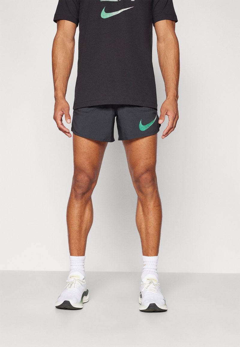 Homme portant un short noir Nike et un t-shirt noir Nike avec des logos Swoosh verts, des baskets blanches et des chaussettes blanches mi-mollet, debout devant un fond uni.