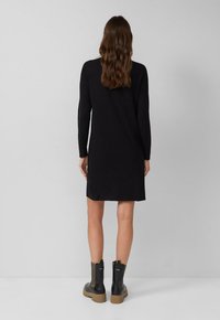 Eine Frau mit welligem braunem Haar steht vor einer grauen Wand und trägt ein schwarzes Langarmkleid sowie schwarze Stiefeletten mit beigen Sohlen.