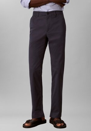 Férfi sötétszürke, egyenes szabású nadrágban és fekete csatos szandálban, karját keresztbe téve, világosszürke, egyszerű háttér előtt.