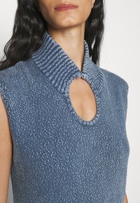 Femme portant un haut en maille bleu sans manches avec un col montant côtelé et une découpe en forme de goutte à l'encolure, dévoilant le haut de la poitrine et le cou.