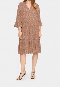 Robe à motif marron avec un décolleté en V, des manches évasées et une jupe à volants. Texture satinée, portée avec des sandales à talons aiguilles avec des lanières.