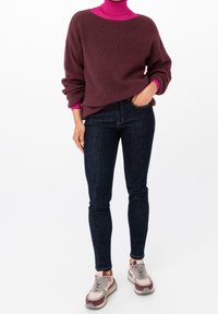 Suéter marrón con canalé sobre un cuello alto fucsia, combinado con jeans ajustados azul oscuro y zapatillas multicolores.