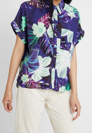 Kortærmet skjorte med knapper og tropisk blomsterprint i lilla, blågrøn og hvid, båret med cremefarvede bukser.