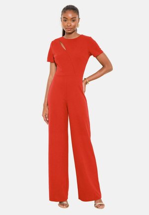 Woman wearing a red short-sleeve jumpsuit with wide legs and a small chest cutout, accessorized with gold earrings, bracelet, and open-toe sandals.