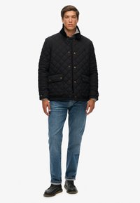Czarna pikowana kurtka z wysokim kołnierzem, zapinana na guziki, z kieszeniami z przodu. Połączona z niebieskimi dżinsami i czarnymi butami. Minimalistyczny design.