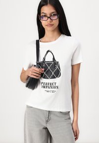 Hvit bomull t-skjorte med grafikk av en svart håndveske og teksten "PERFEKT FØLGE." Grå jeans og en liten svart crossover-veske.