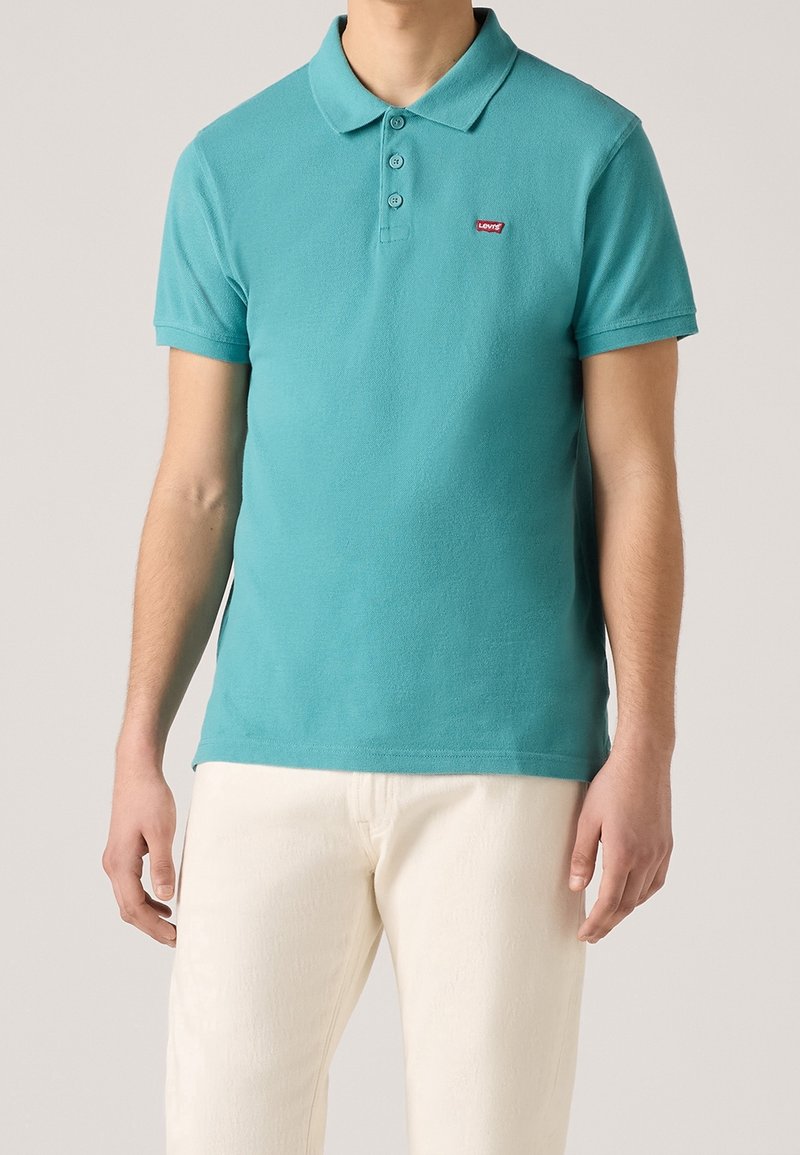 Homme portant un polo manches courtes couleur sarcelle avec trois boutons et un petit logo Levi’s rouge, associé à un pantalon blanc cassé.
