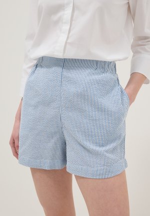 Femme portant une chemise blanche et un short rayé vertical bleu et blanc avec des poches, debout avec une main dans la poche.