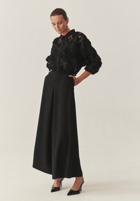 Blusa nera con ricami floreali trasparenti, abbinata a pantaloni neri a gamba larga a vita alta e tacchi a punta neri. Accessori semplici.