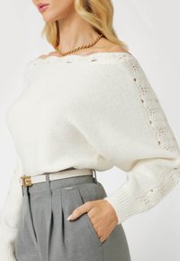 Weißer, schulterfreier Strickpullover mit Spitzen-Details, kombiniert mit hochtaillierten grauen, maßgeschneiderten Hosen und einem weißen Gürtel mit goldenen Beschlägen.