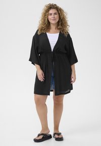 Musta kevytrakenteinen kimono-tyylinen ulkoasu, jossa on kiristettävä vyötärö, leveät hihat ja rento malli. Yhdistetty valkoiseen puseroon ja farkkushortseihin.
