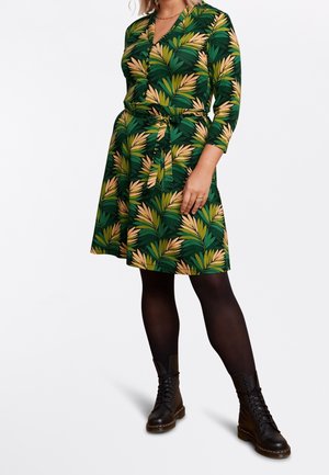 Frau trägt ein knielanges grünes Kleid mit gelbem und grünem Blattmuster, schwarze Strumpfhosen und schwarze Schnürstiefel.