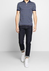 Polo tričko s námořnickými a bílými pruhy s límečkem, kombinované s tmavomodrými džíny a bílými teniskami, ukazující neformální, přiléhavý styl.