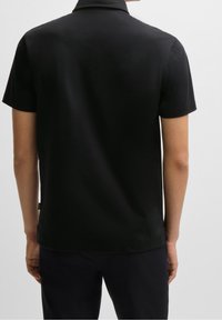 Personne portant un polo noir à manches courtes uni vu de l'arrière contre un fond clair uni.