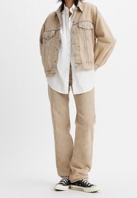 Veste en denim beige avec deux poches poitrine, associée à un pantalon beige taille ample et des baskets en toile noires avec des semelles en caoutchouc blanches.
