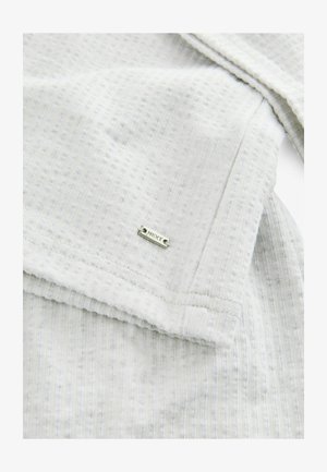 Lys grå stof med et struktureret, ribbet mønster. Har et sølvmærke med påskriften "NEXT" fastgjort nær kanten, hvilket fremhæver et minimalistisk design.