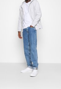 Sweat à capuche gris zippé, t-shirt blanc et jean bleu avec ourlets retroussés ; baskets blanches à surfaces texturées et lacets plats.