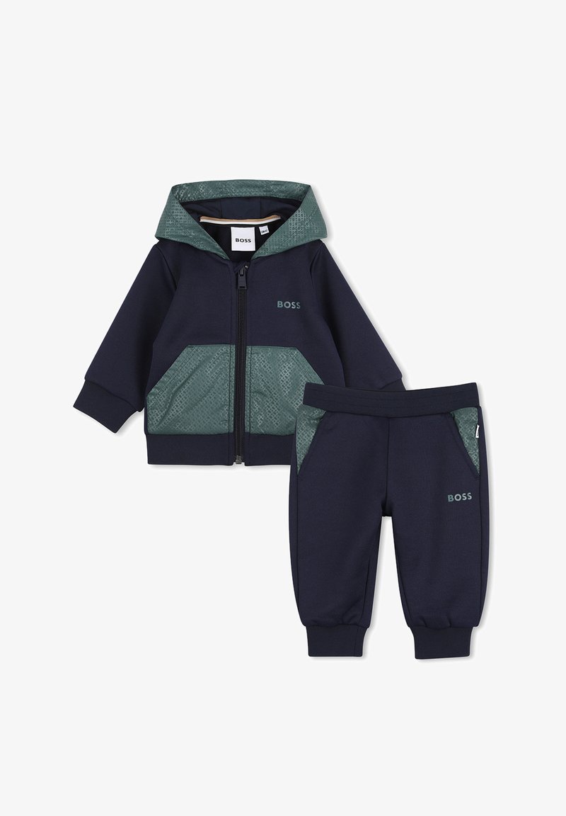 Tuta da bambino blu navy con giacca con cappuccio e zip, dettagli verdi e motivo decorativo, abbinata a pantaloni affusolati coordinati.
