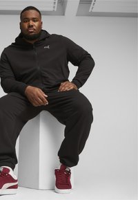 Sort zip-up hættetrøje og matchende joggers i et glat stof, parret med røde og hvide sneakers, siddende på en hvid pedestalsokkel.