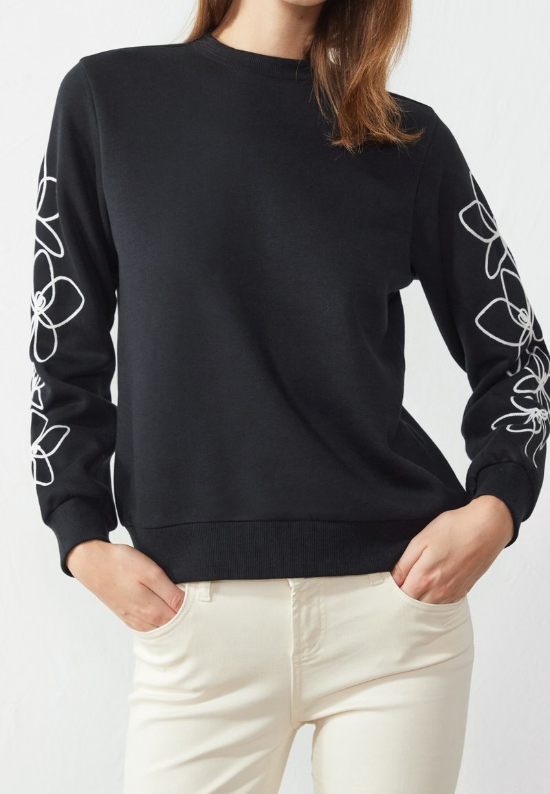 Femme portant un pull noir à manches longues avec des motifs floraux blancs sur les manches et un pantalon blanc cassé, mains dans les poches.