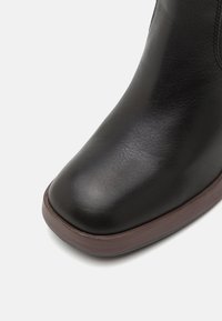 Botină din piele neagră cu vârf rotunjit și textură netedă, având o talpă maro în contrast și un detaliu de cusătură la gleznă.
