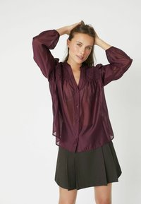 Blusa trasparente bordeaux con colletto cinese, chiusura a bottoni frontale e dettaglio arricciato sulle spalle, abbinata a una gonna plissettata scura.