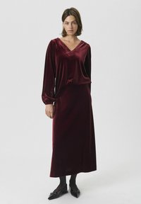 Bordeauxfarbenes Samtkleid mit V-Ausschnitt und langen Ärmeln, das eine lässige Passform und einen subtilen Glanz hat. Kombiniert mit eleganten, spitzen schwarzen Ballerinas.