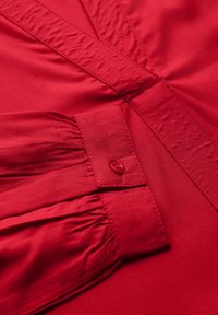 Blusa rossa realizzata in tessuto morbido, con dettaglio arricciato sulla manica e chiusura con bottone. Il design include una linea elegante e sagomata.