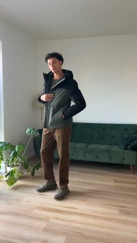 Hættejakke med en mørkegrøn krop og sorte ærmer, lynlås foran, parret med brune cordpants og grå sneakers. Grøn sofa og plante i nærheden.