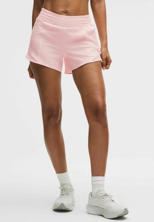 Atleet, kes kannab heledaid roosaid jooksusärke, valgeid sokke ja valgeid jooksujalatseid, seisab ühevärvilise tausta ees.