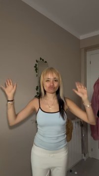 Young woman with blonde hair in a blue tank top raises her hands and makes a playful face in a room with neutral walls and a mirror.