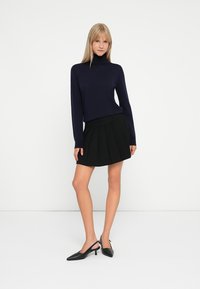 Navy turtleneck sweater, fitted, paired with a black pleated skirt. Model wears black slingback heels. Minimalistic, solid-color outfit.
