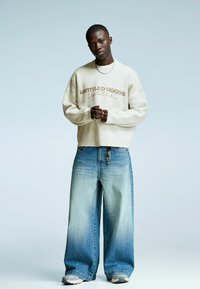 Jonge man in een crèmekleurige trui met de tekst "UNTITLED GOODS", met wijde vervaagde blauwe jeans, een zilveren ketting en grijze sneakers, staand.