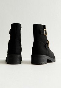 Para czarnych botków za kostkę z grubymi obcasami, złotymi klamrami po bokach oraz teksturowanymi podeszwami, pokazana od tyłu na białej powierzchni.