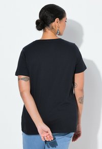 Camiseta negra de manga corta con cuello redondo, textura suave y corte recto, combinada con jeans azul claro; vista trasera muestra tatuajes en los brazos.