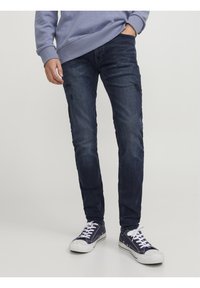 Jeans de mezclilla azul oscuro con corte slim, con detalles desgastados en el frente, complementados con zapatillas blancas y un sudadera azul clara.