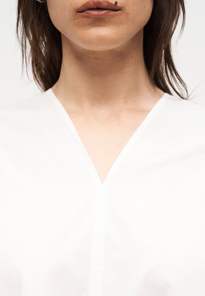Κοντινό πλάνο του κάτω μέρους του προσώπου και του λαιμού μιας γυναίκας, που φοράει λευκό τοπ με V-λαιμόκοψη και έχει σκουλαρίκι στο χείλος στη δεξιά πλευρά.