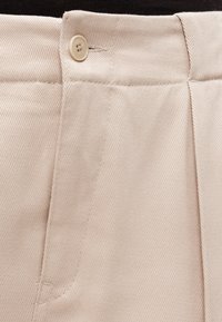 Pantalon en coton beige avec une texture lisse, une fermeture à bouton unique, et un motif en tissage diagonal sur la surface du tissu.