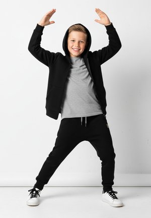 Czarna bluza z kapturem na zamek, z przednimi kieszeniami, w połączeniu z szarą koszulką i czarnymi dresami. Białe trampki z czarnymi sznurowadłami. Prosty i wygodny design.