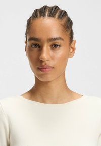 Model wearing a fitted, cream-colored top with a round neckline. Hair is styled in tight braids. Skin tone is medium with a smooth texture.