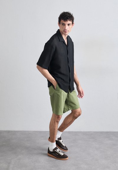 Chemise noire en tissu tissé à manches courtes avec un design à boutons, assortie à un short vert. Des baskets marron et des chaussettes blanches complètent la tenue.